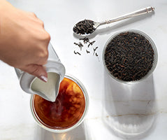 Person pouring tea into a cup with loose tea leaves and a scoop on a white background