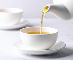 White teacup being filled with tea from a white pitcher on a white surface.